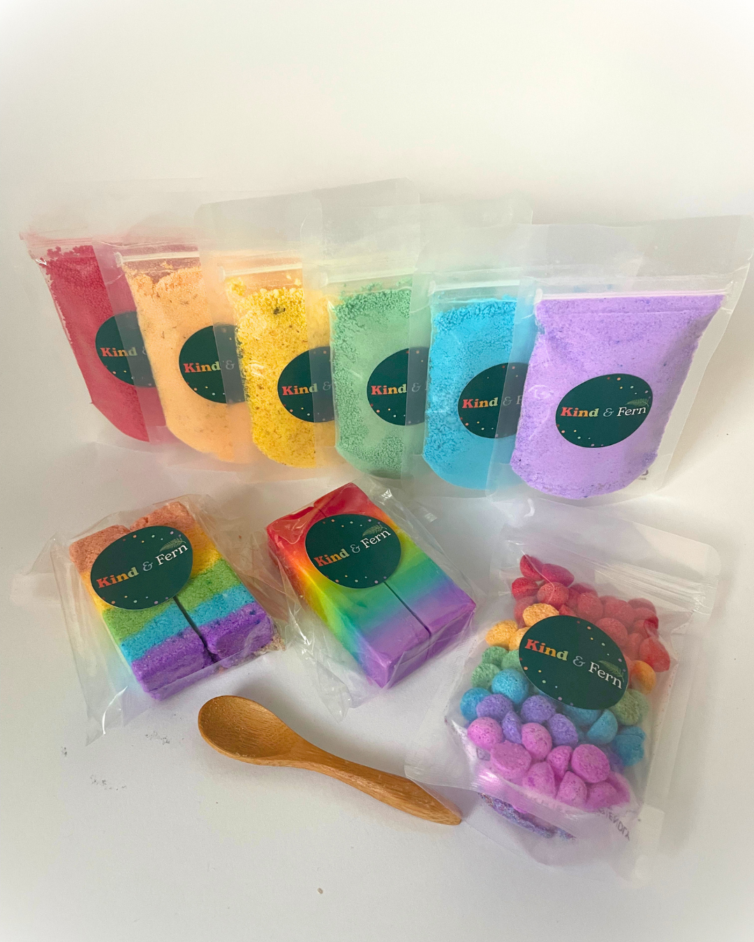 Flat-lay of the Kind & Fern Rainbow box: six colourful bath dust pouches, two rainbow dust bars, rainbow soap, bath teenies and a wooden scoop