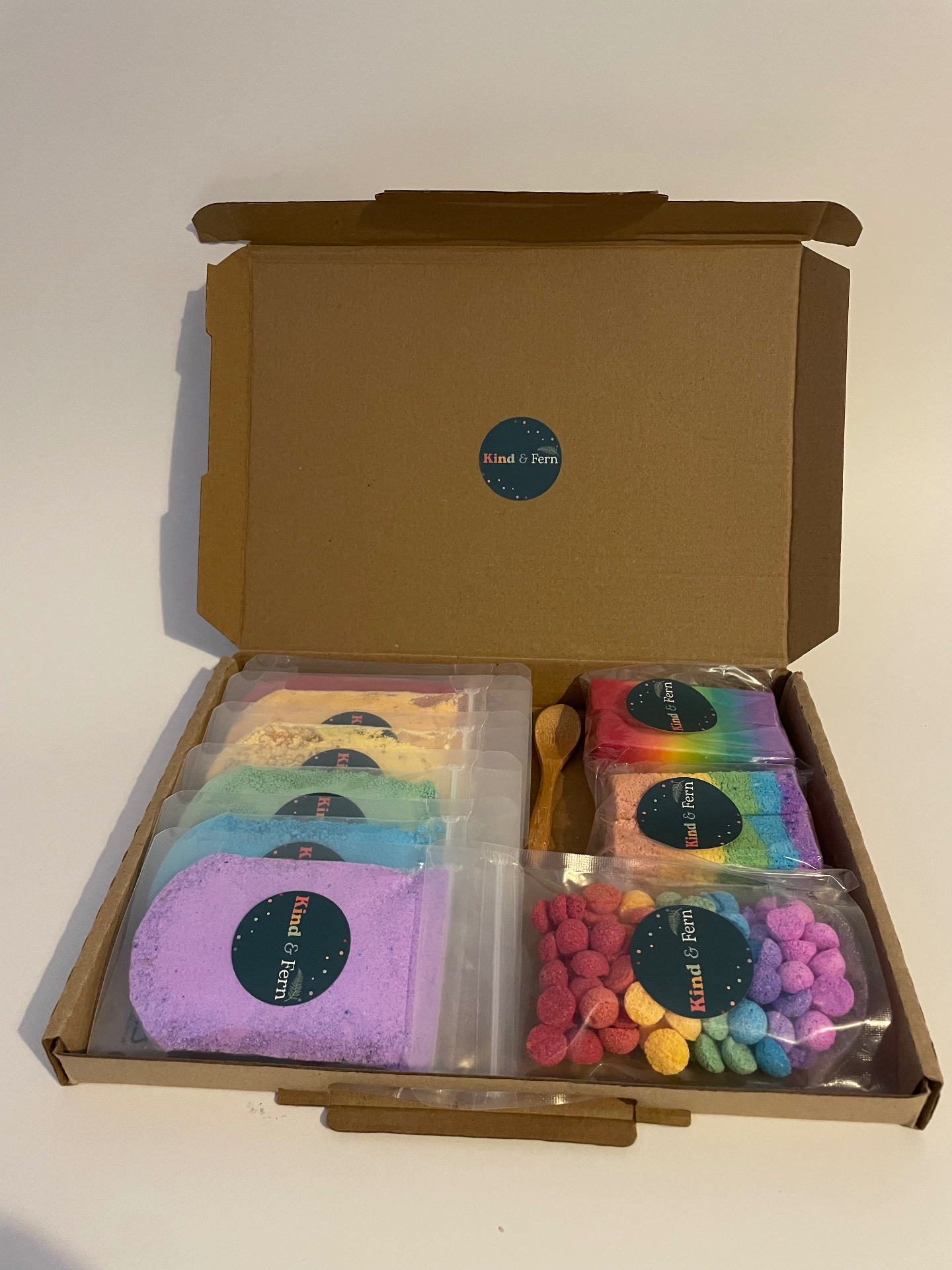 An open Kind & Fern Rainbow letterbox bath box packed with rainbow bath dust pouches, fizz bars, soap, teenies and a wooden scoop