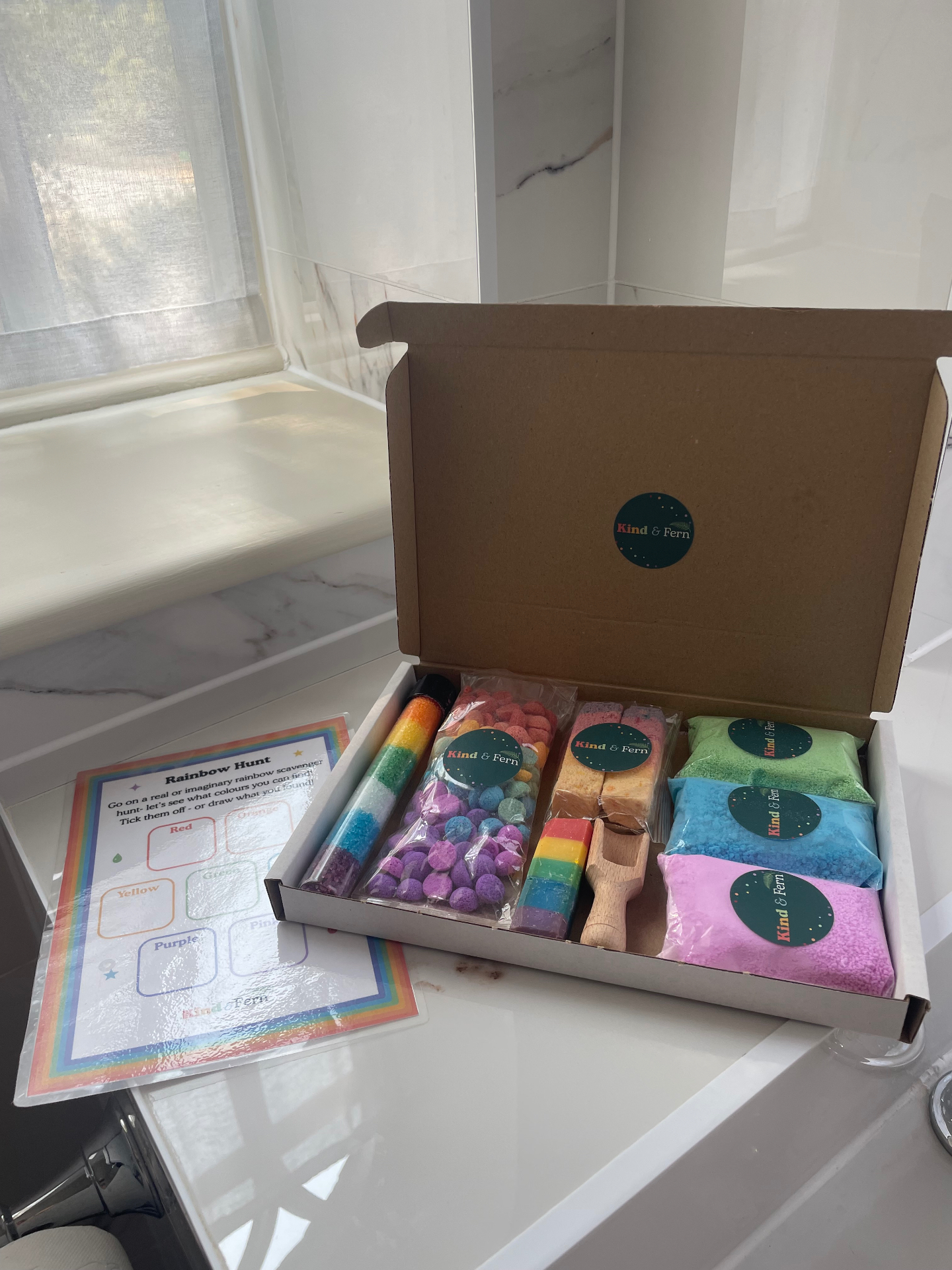 A Kind & Fern Splash Edit bath box open on the side of a bath, filled with rainbow bath products and a Rainbow Hunt printable